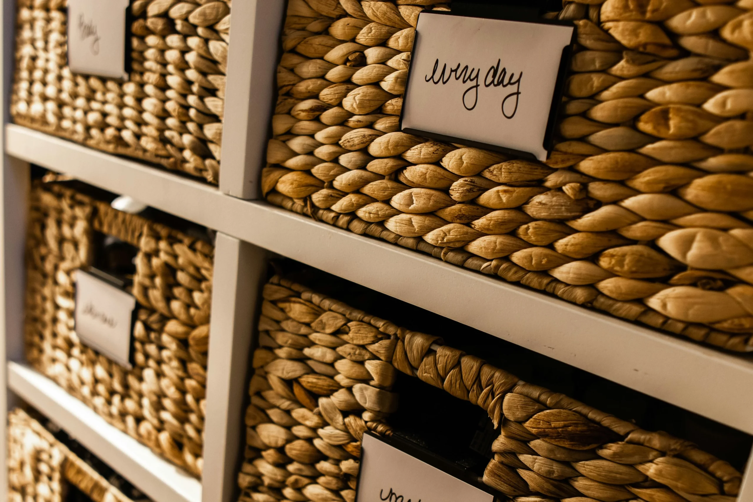 Image of neatly labeled woven storage baskets arranged on white shelving in an organized home storage system