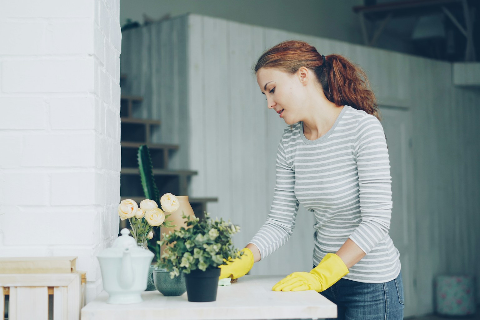 Cleaner in a striped shirt and yellow gloves wiping down a table in a bright home
